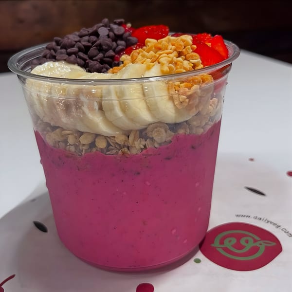 A vibrant pink Daily Veg smoothie bowl with bananas, granola, chocolate chips, strawberries, and peanuts.
