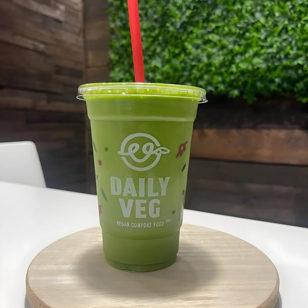 A Daily Veg green smoothie with a red straw on a wooden board, against a green plant wall.